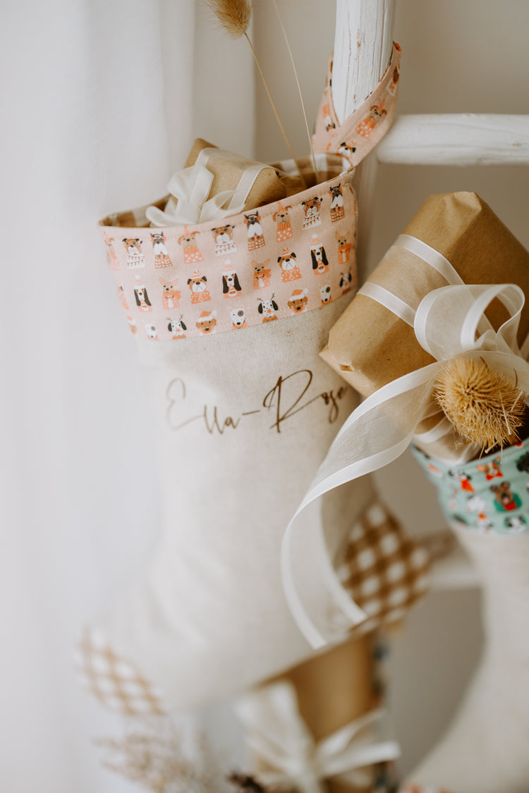 Close-up of handmade personalised girls Christmas stocking in beige fabric with caramel gingham detail and pink cuff featuring festive puppy dog print, customised with name Ella-Raye.