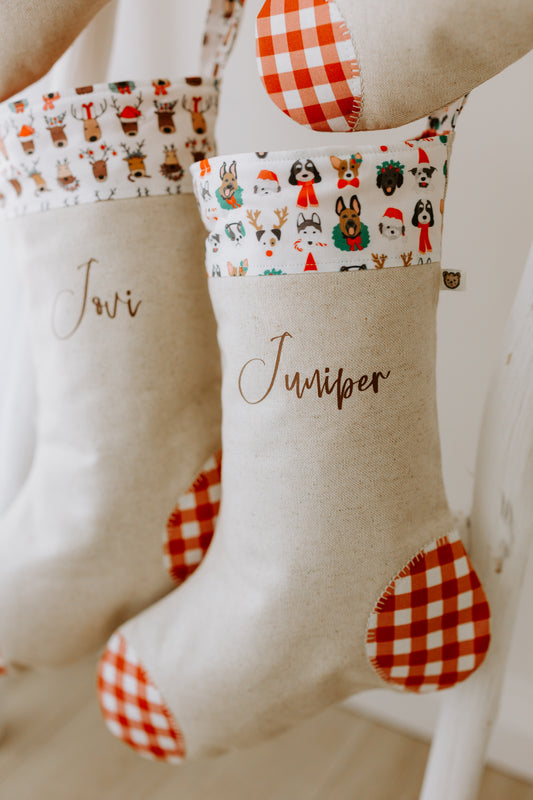 Handmade personalised Christmas stocking in natural linen with red gingham heel and toe patches, featuring the name “Juniper” in brown script. The cuff fabric is decorated with festive puppy dog illustrations — including dogs in Santa hats, Christmas jumpers, and reindeer antlers — making it the perfect stocking for pet lovers or a special Christmas stocking for dogs. A fun and unique holiday decoration for families in Australia who want to include their pets in the festive season.