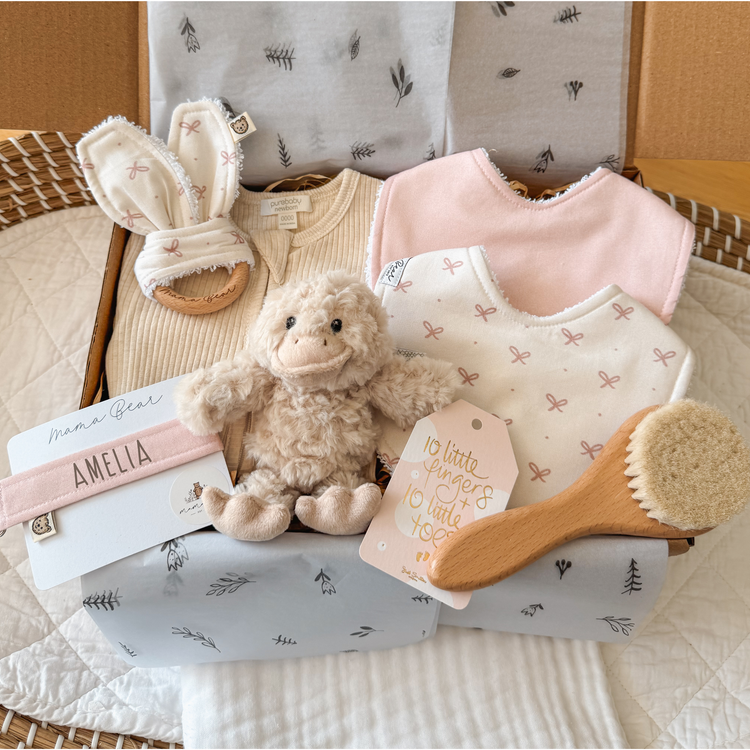 A beautifully arranged newborn baby girl gift hamper displayed in a basket. The hamper includes a soft beige duck snuggly, two handmade bibs in soft pink tones with bow patterns, a personalized pink dummy clip with the name “Amelia,” a natural wooden teething ring with bunny ear fabric, a soft bristle hairbrush, and a neutral-toned zipsuit. The background features a cozy woven basket and soft linens, with a delicate “10 little fingers, 10 little toes” gift tag adding an extra sentimental touch. The presenta