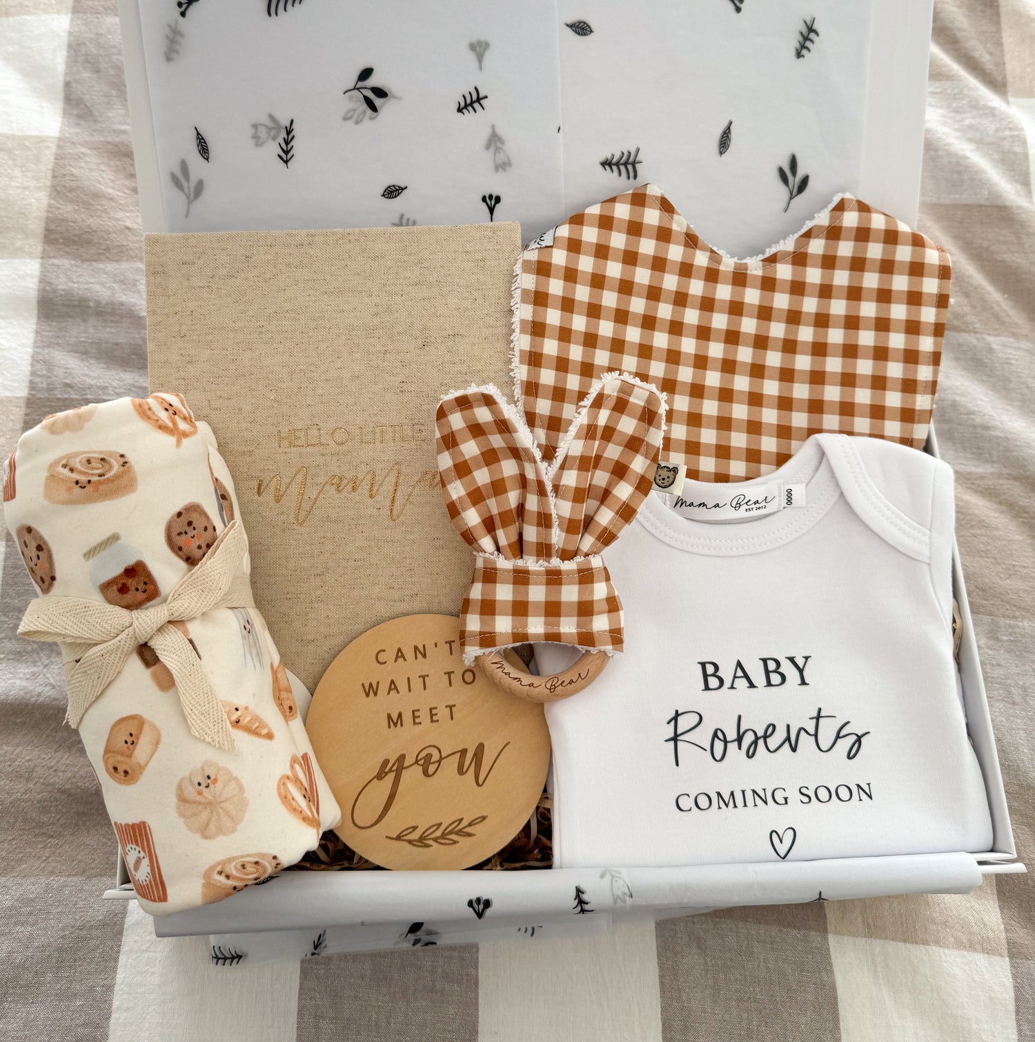 A special gift box for a pregnant Mother to be. Featuring a personalised baby onesie with 'Baby Roberts Coming soon'. A ochre mustard baby bib and matching teething ring. A beige linen pregnancy journal, a wooden plaque that says 'cant wait to meet you' and a soft jersey swaddle with a white background with small illustrations of bakery items such as cookies, scrolls, bread and coffee on it. Packaged beautifully inside a white magnetic close gift box.