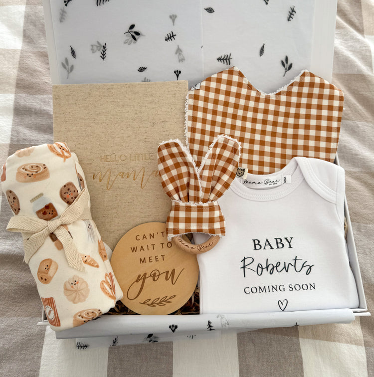 A special gift box for a pregnant Mother to be. Featuring a personalised baby onesie with 'Baby Roberts Coming soon'. A ochre mustard baby bib and matching teething ring. A beige linen pregnancy journal, a wooden plaque that says 'cant wait to meet you' and a soft jersey swaddle with a white background with small illustrations of bakery items such as cookies, scrolls, bread and coffee on it. Packaged beautifully inside a white magnetic close gift box.