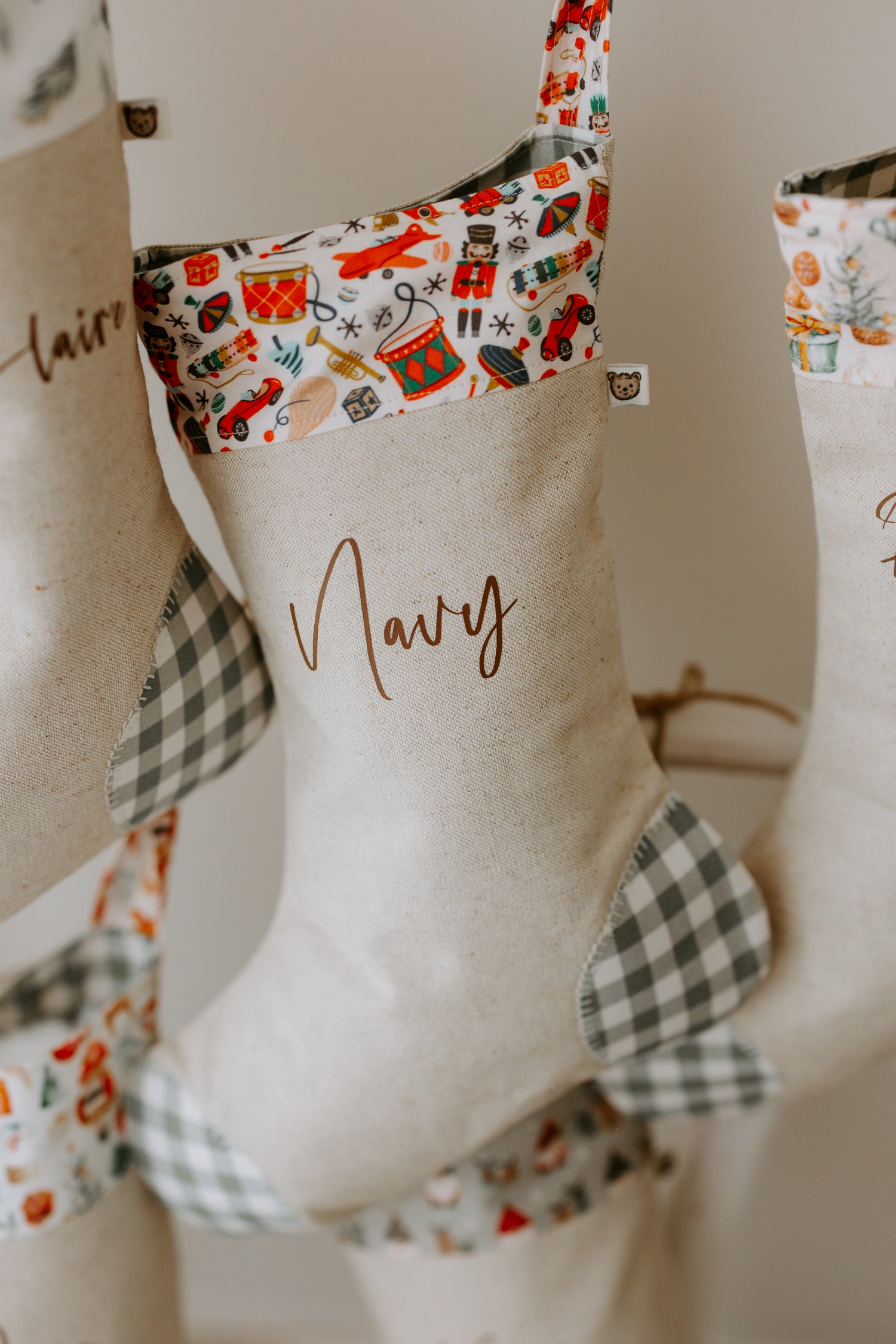 Personalised Christmas stocking in natural beige linen with the name “Navy” in elegant script. The cuff showcases a bright vintage-style Christmas toy print with nutcrackers, toy drums, rocking horses, and Santa figures in festive red and green tones. Grey gingham fabric is used on the heel and toe patches, giving the stocking a timeless handmade look that suits both kids and family holiday décor.