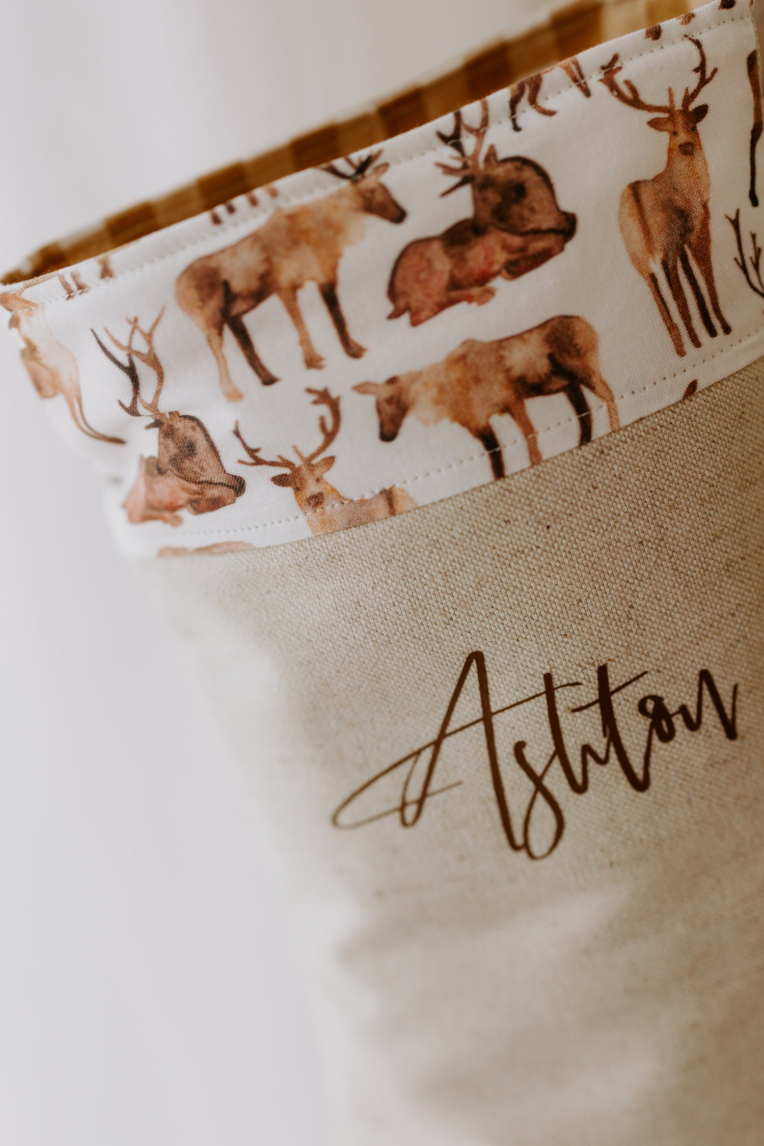 Close-up of a personalised Christmas stocking made from natural linen fabric, featuring the name “Ashton” in brown script. The stocking has a cuff decorated with a woodland reindeer print in soft watercolour tones of brown and beige, giving it a rustic, nature-inspired Christmas look. A unique handmade stocking perfect for kids, with a woodland animal theme that complements rustic holiday décor.