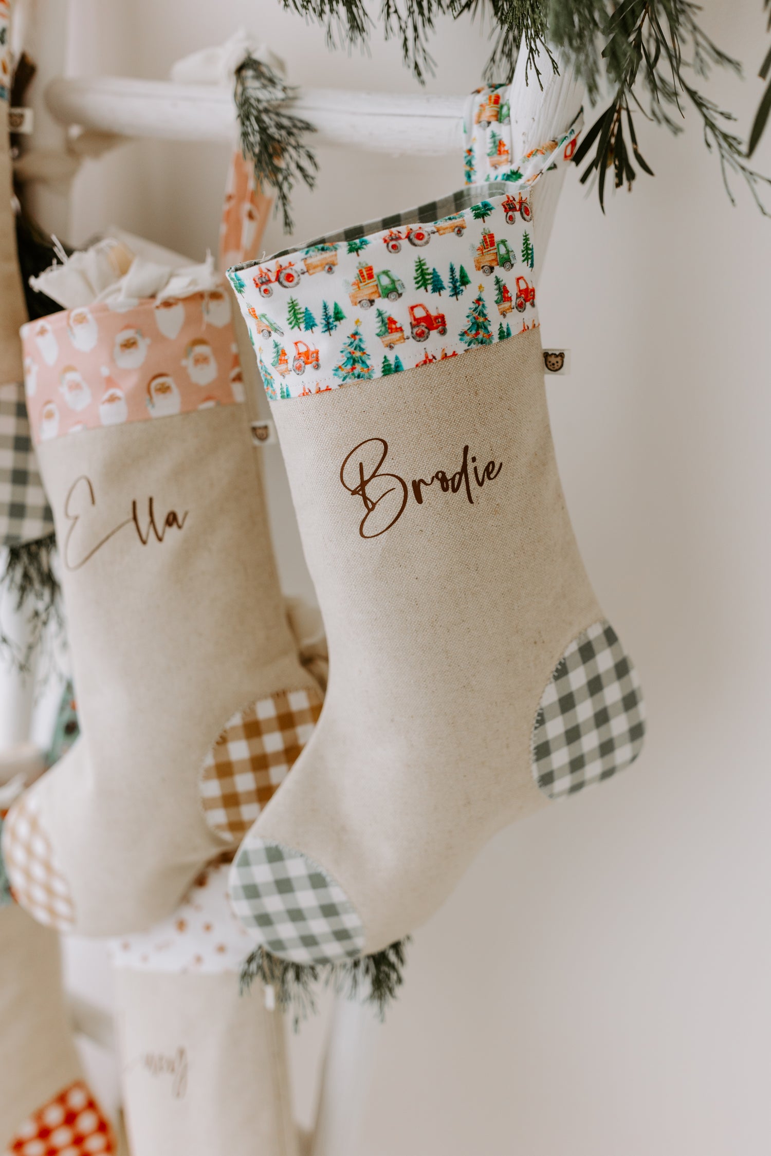 Close-up of two personalised Christmas stockings hanging from a rustic white ladder, one with “Ella” in script font and peach Santa print cuff, the other with “Brodie” and a festive farm design featuring red tractors, Christmas trees, and toy trucks. Both stockings are handmade from natural linen with gingham patchwork heels and toes, styled with sprigs of greenery for a cosy farmhouse holiday look.