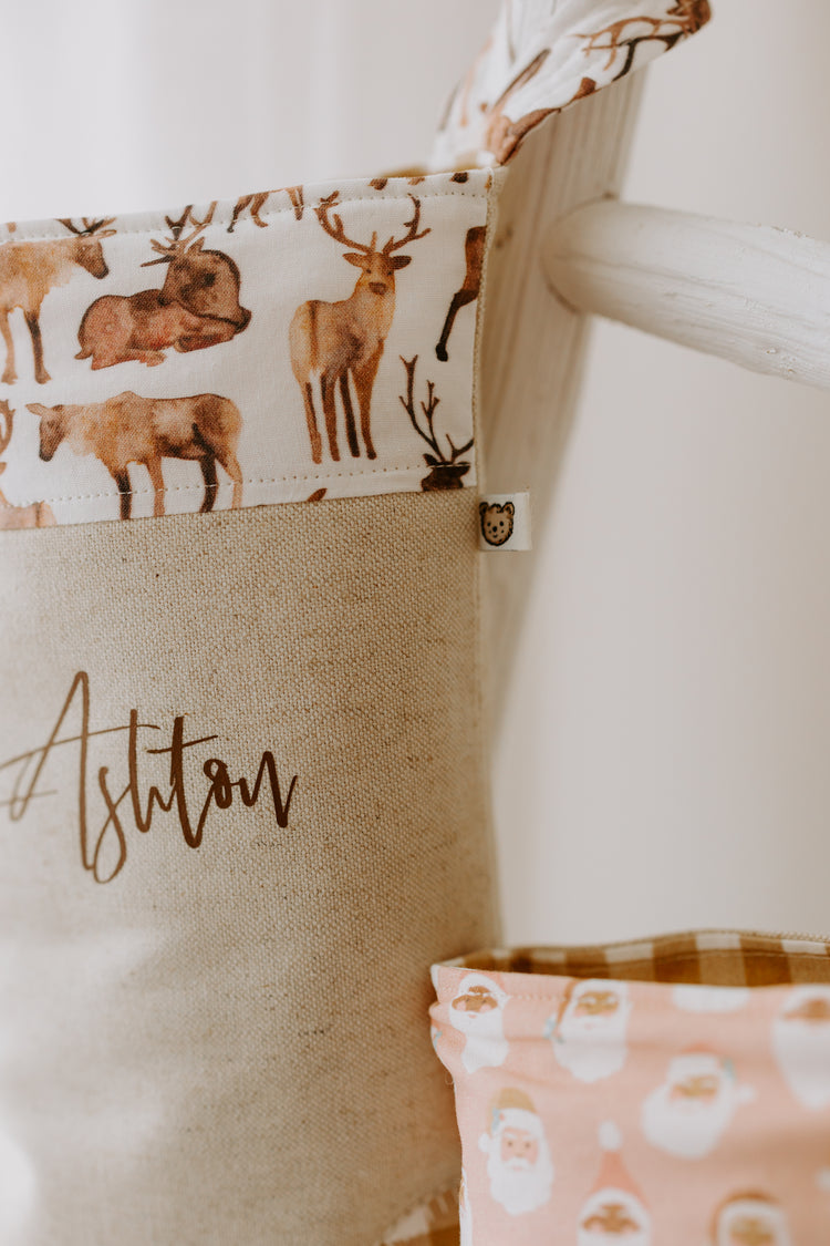 A unisex Christmas stocking suitable for the whole family. Stocking is in a beige linen, with a brown script font to personalise name. The trim has hand drawn reindeers on a white background. A very minimal design.
