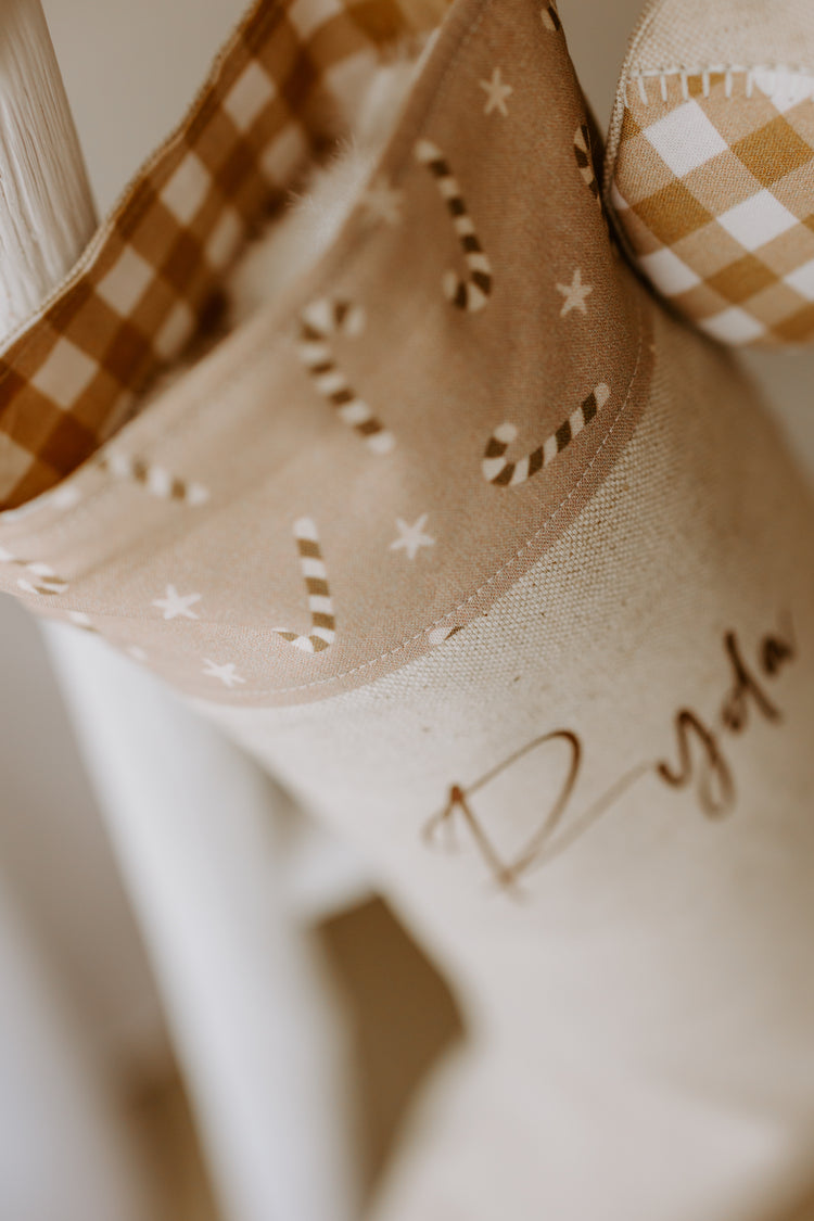 Close-up of handmade personalised Christmas stocking in beige fabric with caramel gingham trim and candy cane print cuff, customised with name Ryda.
