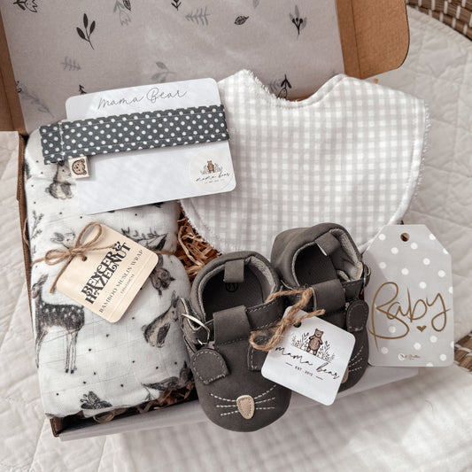 Baby Gift set in grey tones featuring a woodland swaddle (white background, deers, bunny, owls). A light grey gingham baby bib, a dark grey spot dummy clip and a pair of dark grey mouse baby shoes.