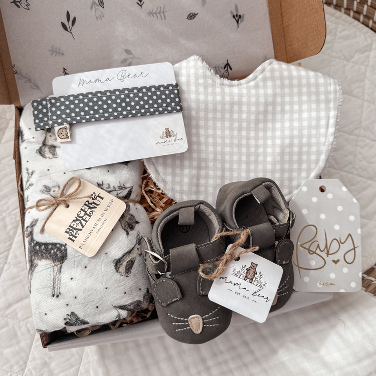 Baby Gift set in grey tones featuring a woodland swaddle (white background, deers, bunny, owls). A light grey gingham baby bib, a dark grey spot dummy clip and a pair of dark grey mouse baby shoes.