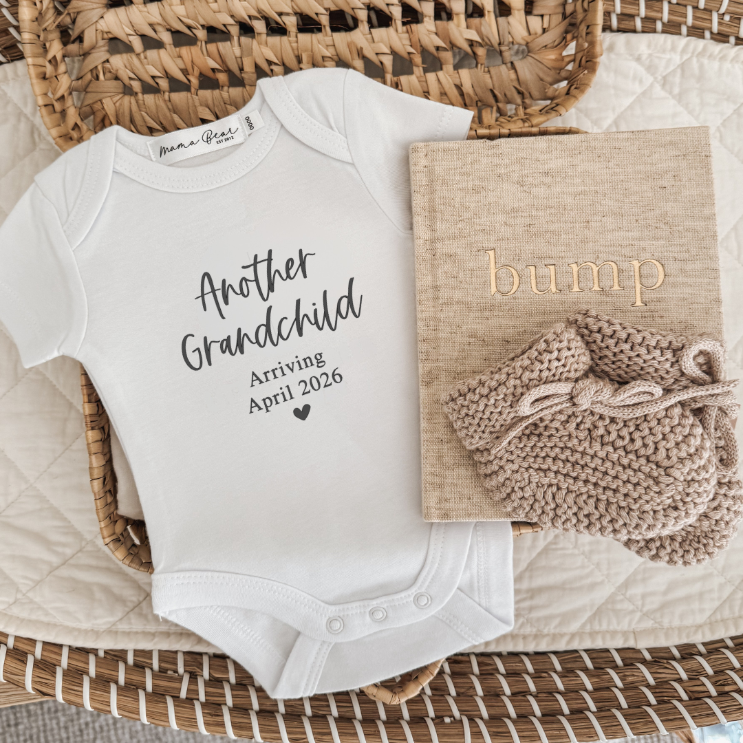 White baby onesie with text 'another grandchild' (script font), 'Arriving April 2026' in times roman. The onesie is laying in a woven basket with some beige baby booties and a linen beige bump journal.