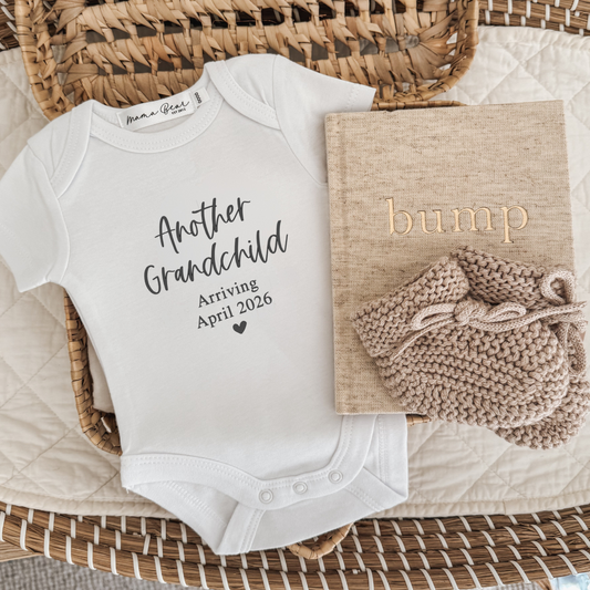 White baby onesie with text 'another grandchild' (script font), 'Arriving April 2026' in times roman. The onesie is laying in a woven basket with some beige baby booties and a linen beige bump journal.