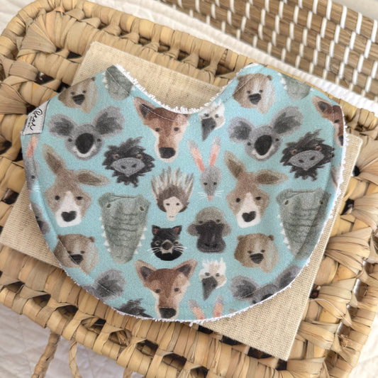 Flat lay baby bib with Australia animal faces illustrated on it. Background is light blue. Some animal faces on the bib are Crocodile, koala, emu , kangaroo , dingo , wombat.
