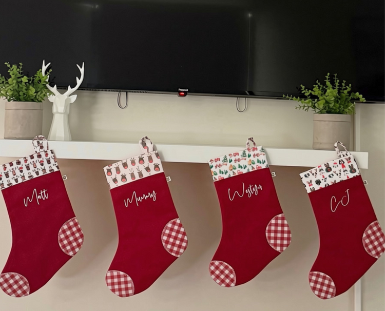 Handmade red and red gingham christmas stockings personalised with names in white script font. The trim at the top of stocking is white with illustrations of christmas reindeers or christmas puppy dogs.