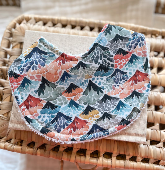 Handmade baby bib with a bold volcanic print featuring erupting mountains in shades of red, orange, mustard, blue, and charcoal. The lava flows and textured details create a fiery, adventurous feel. Bib is rounded in shape with terry towelling backing, shown on a linen cloth and woven basket.