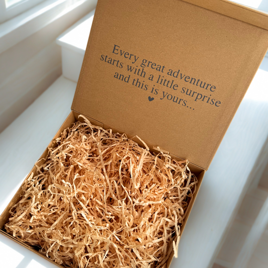 Open kraft magnetic gift box filled with natural shredded paper. The inside of the lid is printed with the message “Every great adventure starts with a little surprise and this is yours…” in black text, creating a warm and sentimental reveal for a pregnancy announcement gift.