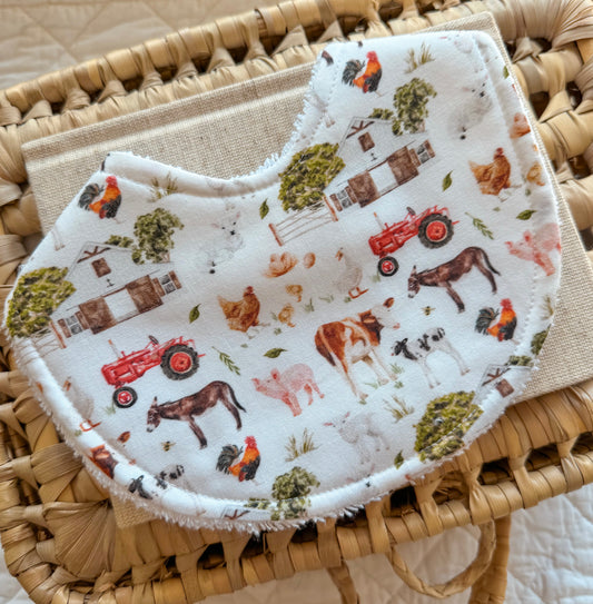 Flat lay of a handmade baby bib with a farm animal print featuring cows, pigs, chickens, donkeys, sheep, trees, barns, and red tractors on a soft white background. Bib is styled in a woven basket.