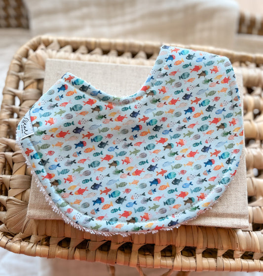 Handmade baby bib featuring a fun underwater pattern with small, colourful fish in shades of orange, teal, green, and blue. The fish are scattered evenly across a pale blue background. The bib is rounded with terry towelling backing, shown on a neutral cloth and woven basket.
