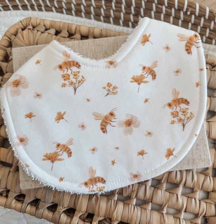 Baby bib with little bees and flowers scattered across it with a white / pale blush background.