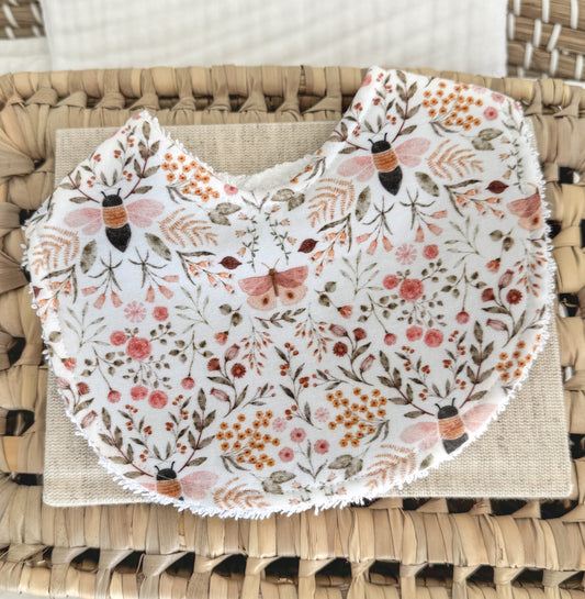 Handmade baby bib with a detailed nature print including honeybees, butterflies, and tiny floral elements in soft peach, rust, and muted green on white cotton, contoured for newborn comfort and ideal for gifting.