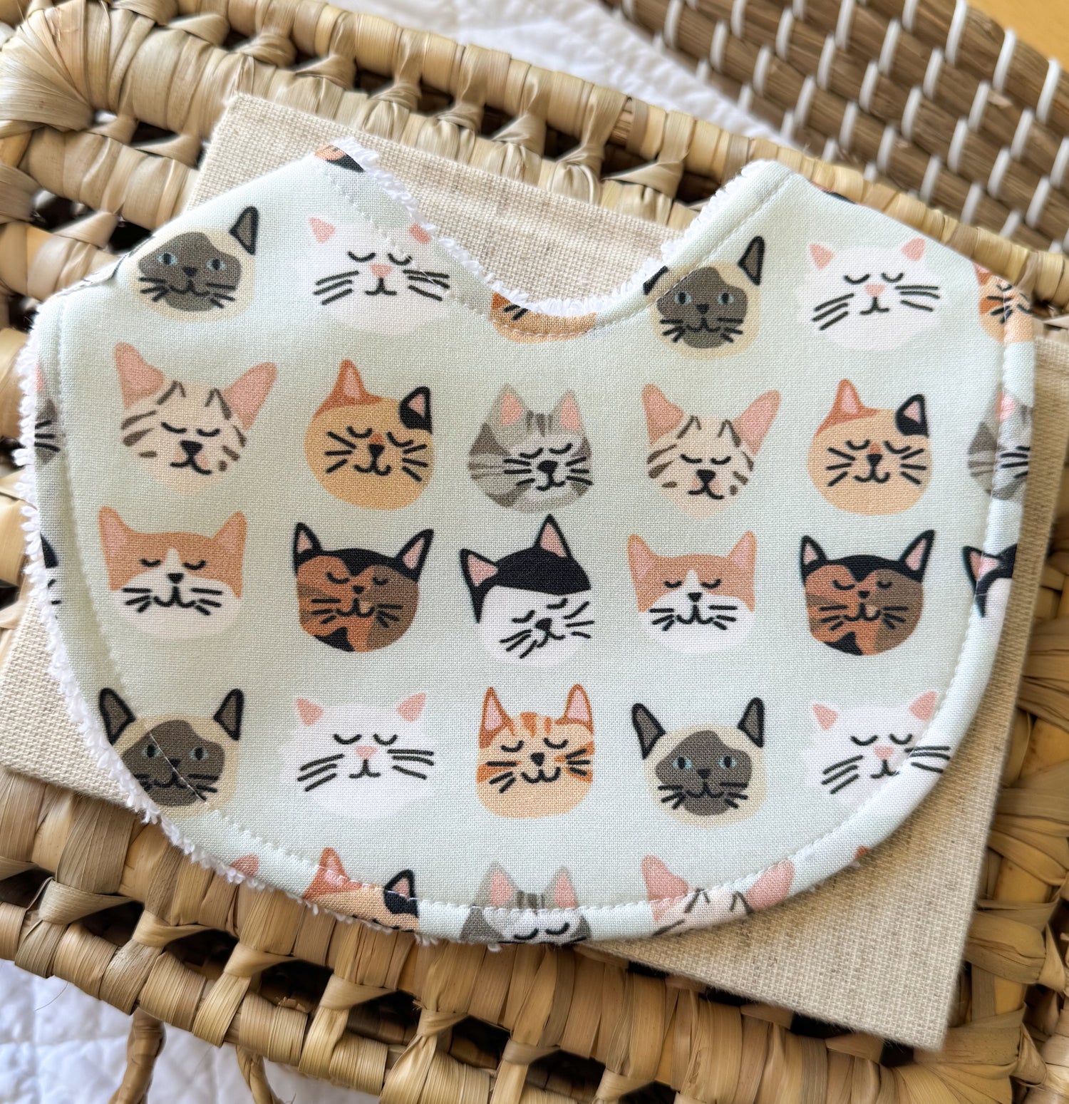 Handmade baby bib with a very pale green background with little cat face illustrations. 