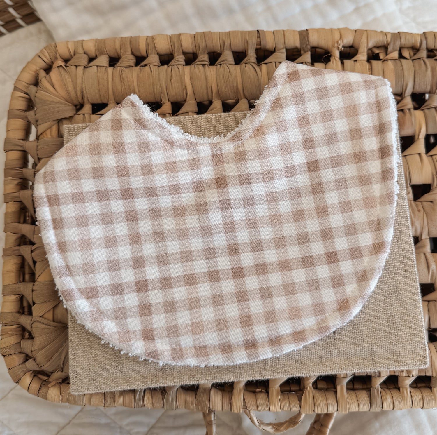 Neutral-toned handmade baby bib in a soft stone gingham print, placed on a beige linen cloth over a woven basket. The bib features curved edges and terry towelling backing, ideal for teething and dribble.
