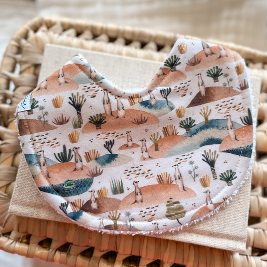 Baby bib with a hand-illustrated desert landscape featuring standing meerkats, trees, and earthy terrain, backed with absorbent terry towelling for dribble and teething; handmade in Australia.