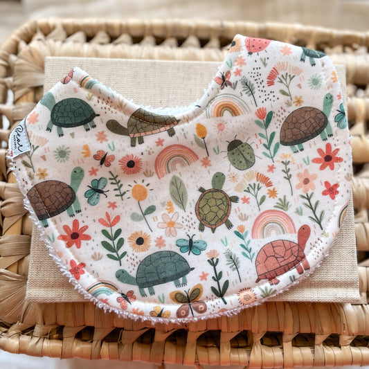 Handmade baby bib with a whimsical nature print featuring turtles, butterflies, rainbows, and wildflowers in soft earthy tones. The curved bib is backed with terry towelling and photographed on a beige linen cloth and woven basket.
