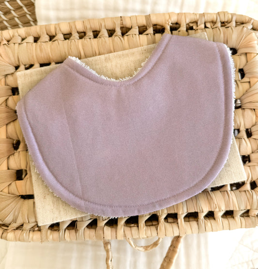 Flat lay of handmade baby bib in solid mauve purple lying on a wicker basket.