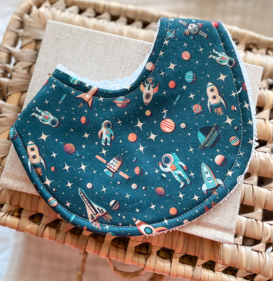 Handmade baby bib with astronauts, rockets, planets, and stars on a dark blue space-themed background; backed with absorbent terry towelling, perfect for newborns and teething babies.