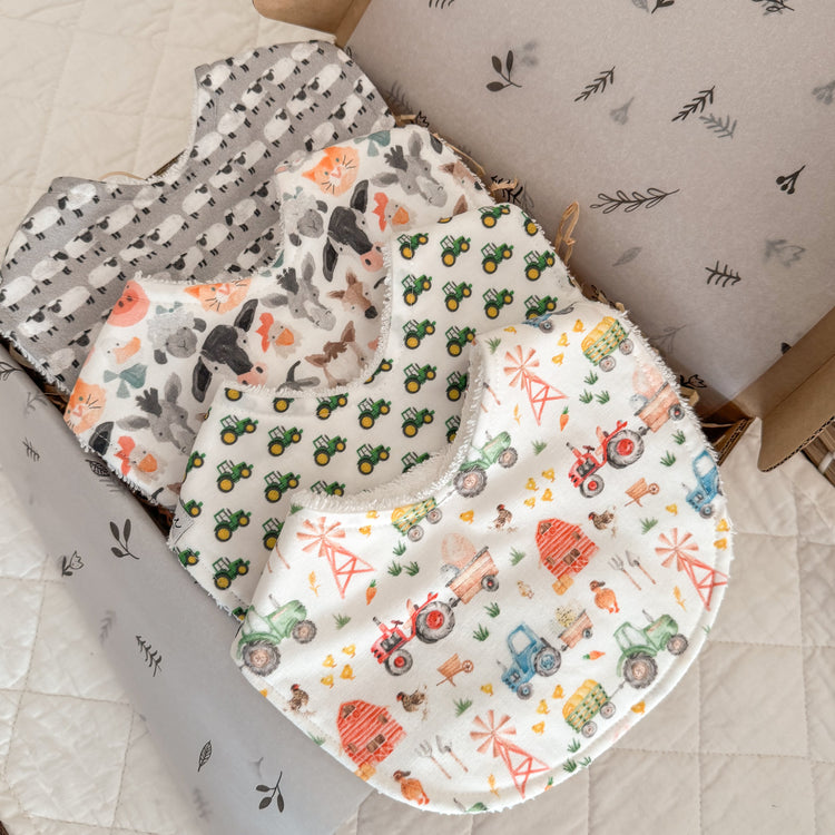 A set of handmade baby bibs with a farm animal and tractor theme, displayed in an open gift box.