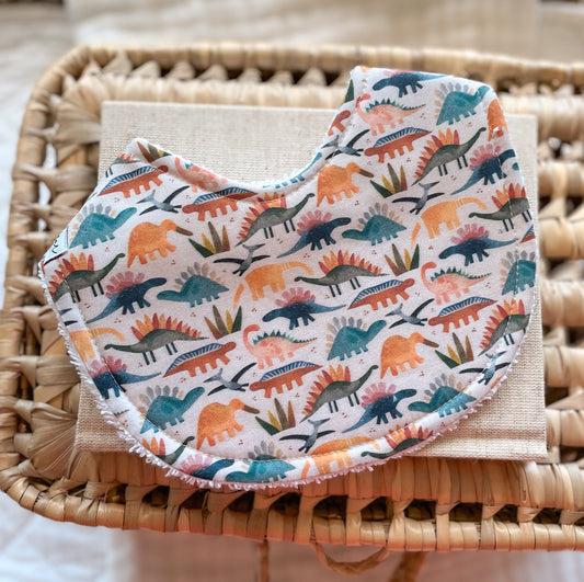 Handmade baby bib with a colourful dinosaur print in shades of teal, burnt orange, mustard, and soft grey on a cream background. Features a mix of stegosaurus and other dinosaur shapes with little leaves scattered throughout. Curved shape with terry towelling backing, photographed on a linen cloth and woven basket.