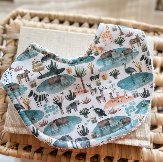 Gender-neutral baby bib with illustrated zebras, giraffes, hippos, birds, and meerkats gathered at desert waterholes; handmade in Australia with soft, absorbent terry towelling backing.