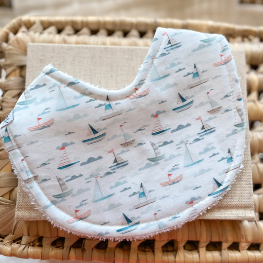 Handmade baby bib featuring a nautical sailing pattern with small boats and yachts in pastel blue, navy, mint, and coral tones. Boats are scattered across a cream background with soft grey clouds. The bib has a curved shape and terry towelling backing, shown on a linen cloth and woven basket.