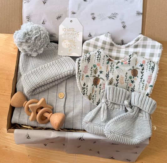Flat lay of a newborn baby boy gift set featuring a pale blue knit beanie, matching booties, a wooden rattle, soft blue romper, two cotton bibs with gingham and turtle prints, and a “10 little fingers 10 little toes” gift tag, all beautifully arranged in a gift box with tissue paper.