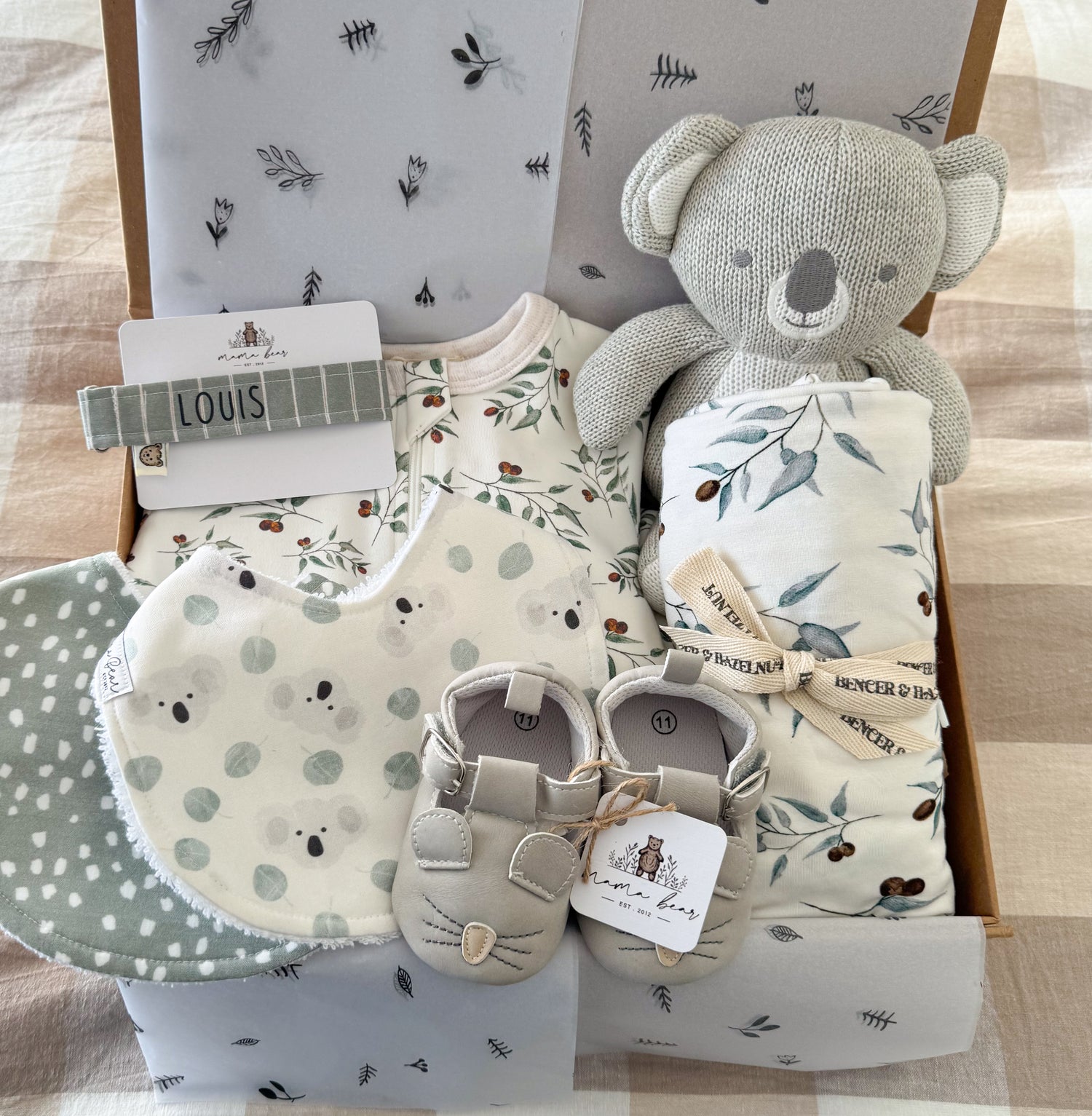 Green eucalyptus theme baby gift box featuring a zip suit and swaddle in green leaves, a cuddly koala toy, a personalised dummy clip made out of sage green stripe material, two baby bibs, one with mini koala faces and the other with a green background and white spots and a pair of light grey mouse baby shoes