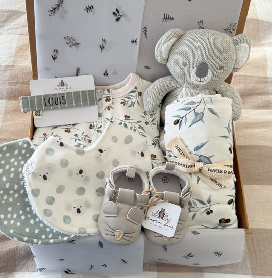 Green eucalyptus theme baby gift box featuring a zip suit and swaddle in green leaves, a cuddly koala toy, a personalised dummy clip made out of sage green stripe material, two baby bibs, one with mini koala faces and the other with a green background and white spots and a pair of light grey mouse baby shoes