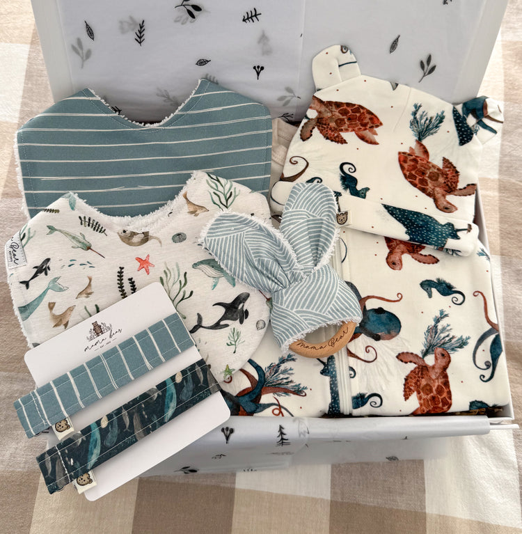Luxury baby boy gift idea featuring an ocean them zipsuit and matching beanie with sea turtles, octopus, sting rays and seahorses in shades of blue and rust orange on a white background. Two handmade bibs, one is deep blue with fine white horizontal stripes in white. The other bib has illustrations of sea creatures such as whales, seals, coral, fish. Two matching dummy clips and a teething ring in a light blue and white wave like fabric to mimic waves of the ocean. Packaged in a white gift box.