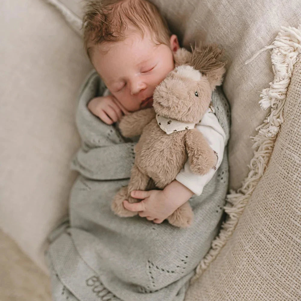 A baby lying on a textured surface holding a plush toy horse rattle.
