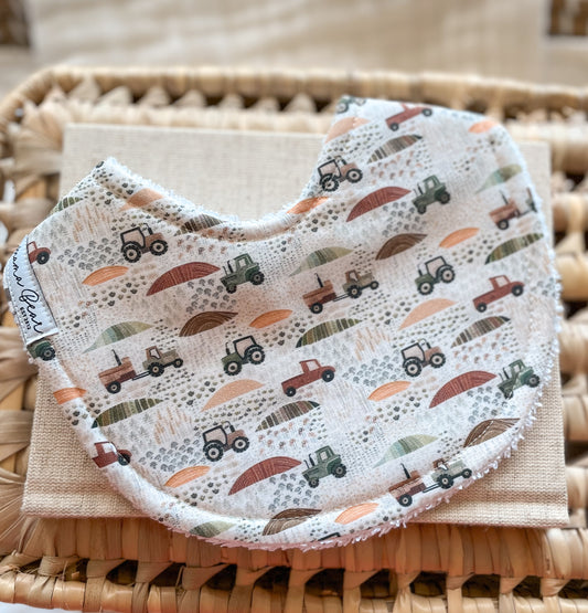 Curved handmade baby bib with a neutral farm print featuring green and brown tractors, red utes, and scattered fields in warm earth tones. The bib is backed with terry towelling and displayed on a beige cloth and woven basket.