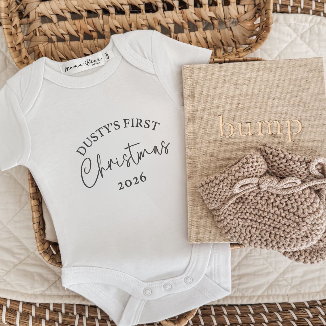 White baby onesie personalised with 'Dusty's first Christmas 2026 in black font. Text is in capital times font and Christmas is in a script font.