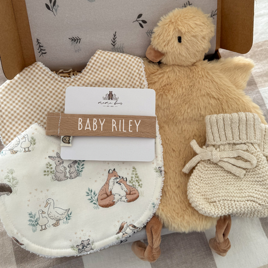 A baby gift set arranged in a kraft box, featuring a cream bib with woodland animal illustrations, a beige gingham bib, a plush yellow duck toy, and cream knit booties, with a light brown personalised dummy clip Baby Riley.