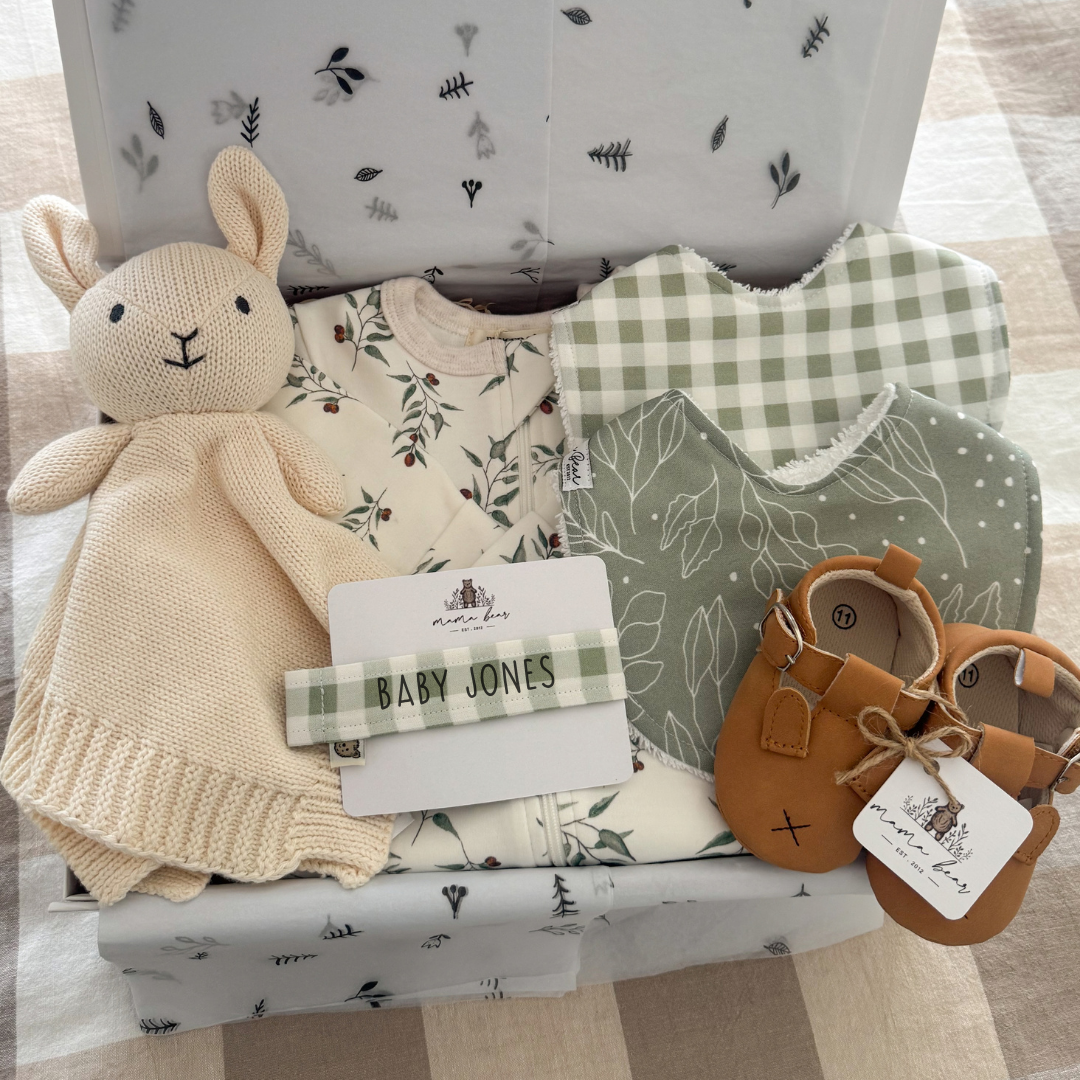 Gender neutral baby gift box with white bunny comforter, baby long sleeve zipsuit with white background and eucalyptus leaves, handmade bibs in green leaf print and sage green gingham check, tan baby bunny shoes and a personalised dummy clip in sage gingham. All packaged inside a white magnetic close Giftbox.
