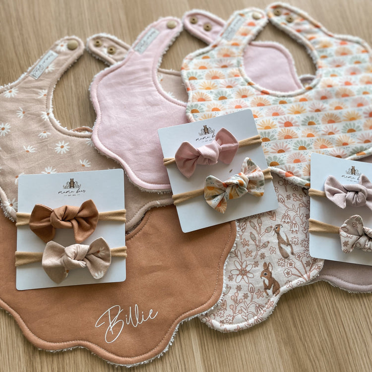 Handmade baby girl bibs in a scollop petal shape. Fabric colours in photo include a solid tan white personalised in white vinyl with 'Billie' script font. A beige daisy bib, solid pink, a pale pink bib with bunnies on it and some matching hair bows.