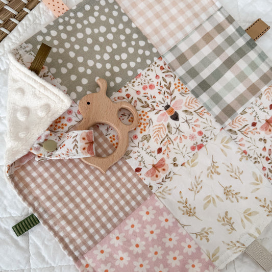 Patchwork quilt style baby taggie with pink florals, polka dot, and checkered patterns. Attached to the taggie is a wooden bee shape. The Blankie includes soft pink, beige and sage green tones.