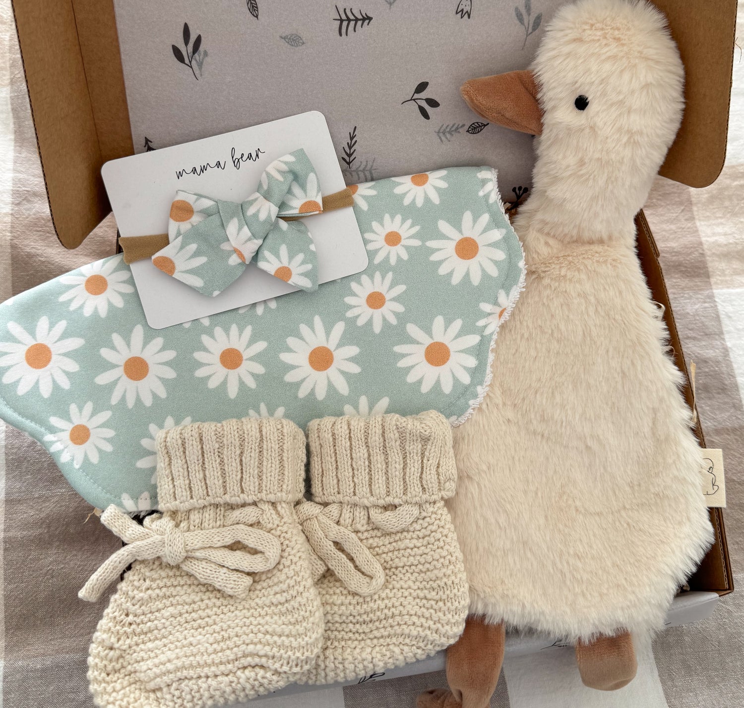 A baby gift set arranged in a kraft box, featuring a plush cream goose toy, a aqua and white daisy-patterned bib with scalloped edges, a matching daisy-patterned baby bow and a pair of cream knit booties.