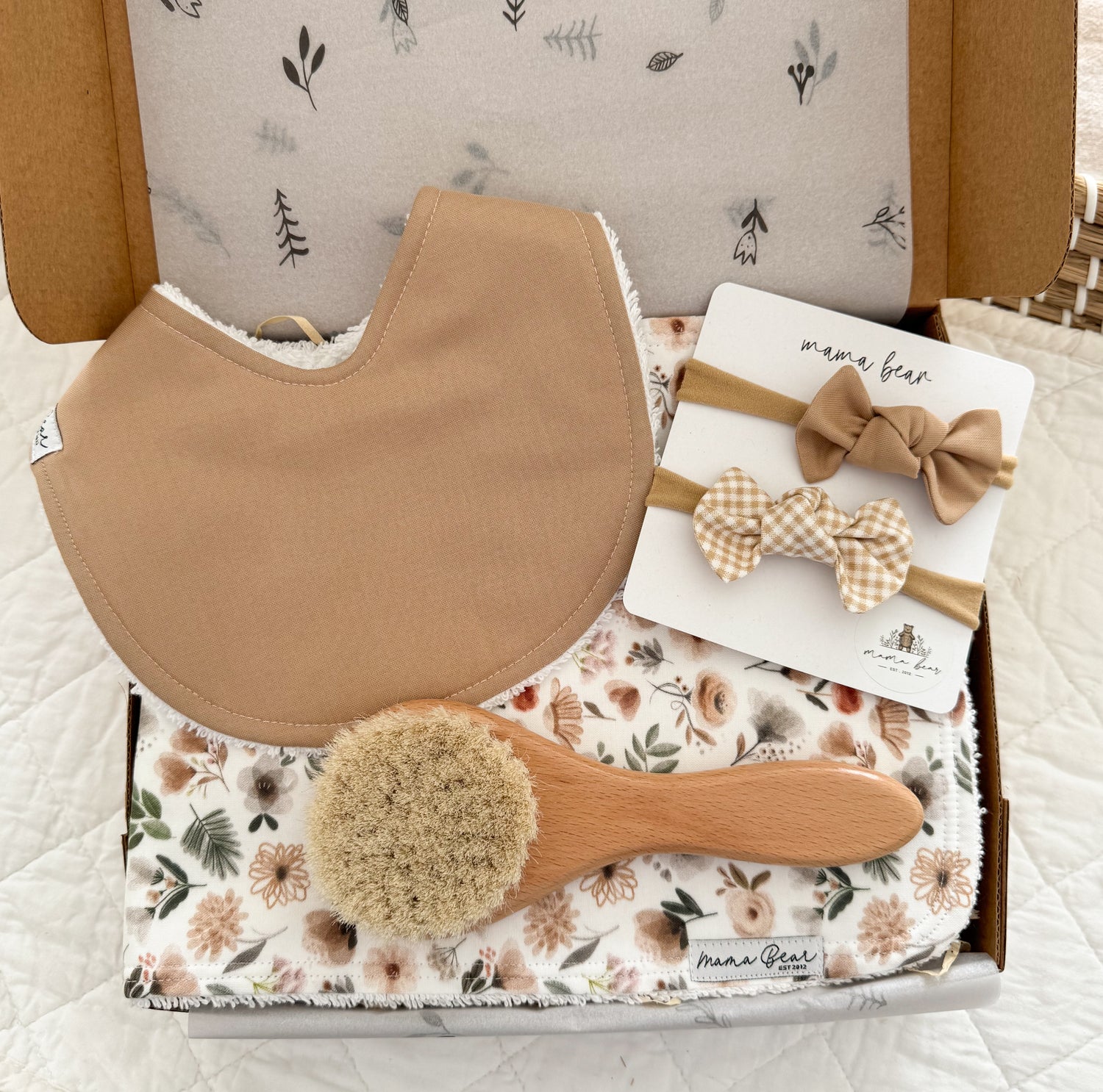 A baby gift set in a kraft box featuring a beige baby bib, a round wooden hairbrush, a woodland theme floral burp cloth in muted beige, sage tones and 2 matching hair bows.
