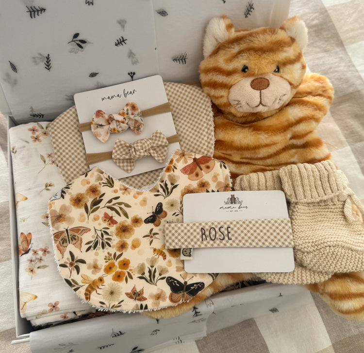 Baby girl gift box featuring a beige gingham baby bib, another handmade bib in beige earthy tones with flowers, butterfly and bees on it, floral & butterfly muslin wrap, beige knitted baby booties, plush ginger cat comforter toy, personalised baby dummy clip, matching floral and gingham hair bows. Packaged inside a large white magnetic closure gift box.