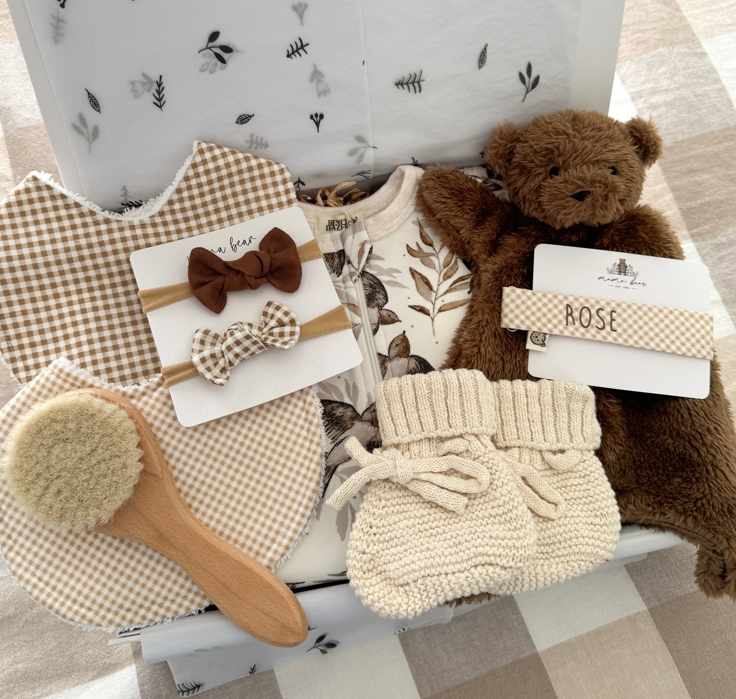 A baby gift set arranged in a white gift box, featuring a brown teddy bear, cream knit booties, two brown and light brown gingham bibs, a wooden baby brush, two baby bow headbands in brown gingham and plain chocolate brown, and a matching gingham dummy clip labeled 'ROSE' in brown vinyl.