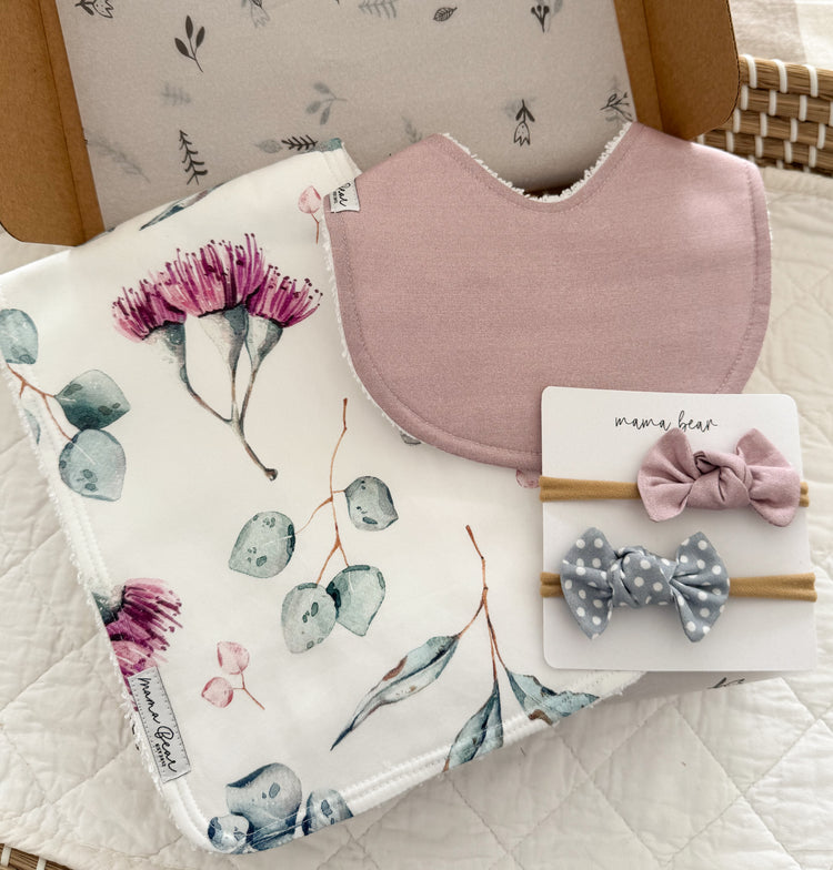 A baby gift set featuring a musk pink bib, a burp cloth with watercolor eucalyptus and pink flower blossom print,  a matching pink hair bow and a gray polka dot hair bow, all arranged in a kraft brown gift box.