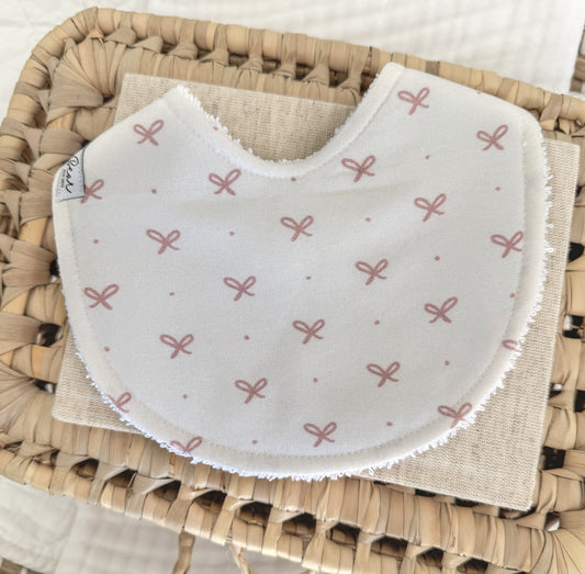 Baby bib for girls with an all-over blush pink bow print on soft white cotton, handmade in a rounded contoured shape for comfort and absorbency.