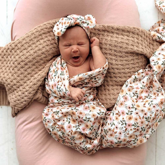 Newborn baby wrapped in a floral blanket with a matching headband, lying on a pink surface.